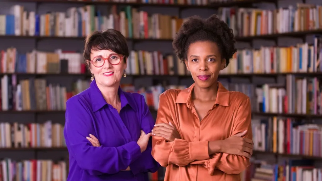 Fotografia delle fondatrici di Bold Stories, Cristina Maurelli e Giuditta Rossi. La foto le ritrae a mezzo busto. Sullo sfondo una libreria. Maurelli è bianca, ha degli occhiali rossi e i capelli corti, indossa una camicia viola ; Rossi è nera, ha i capelli raccolti e indossa una camica sui toni dell'arancione.